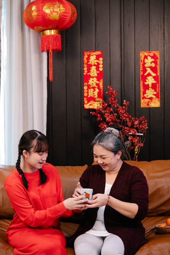 Cheerful ethnic female teenager passing present cup to grandma on couch during New Year holiday at home